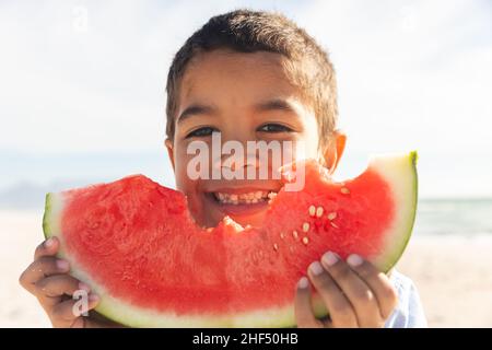 Porträt eines lächelnden niedlichen Biracial Jungen hält große frische Wassermelone Scheibe am Strand an sonnigen Tag Stockfoto