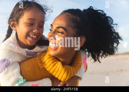 Fröhliche junge Birazialfrau, die ihre Tochter ansieht, während sie während des Sonnenuntergangs ihre Huckepack-Fahrt macht Stockfoto