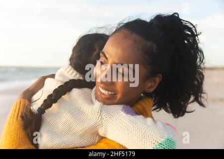 Lächelnde junge Birazialfrau umarmt Tochter, während sie am Strand während des Sonnenuntergangs getragen Stockfoto