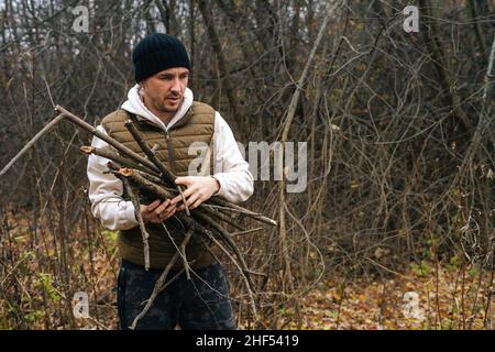 Portrait eines überlebensfähigen Mannes, der warme Kleidung trägt und trockenes Totholz für das Feuer im Wald an einem bewölkten, kalten Tag sammelt. Stockfoto