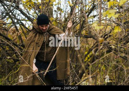 Porträt eines ernsthaften Touristen-Mannes, der im Regenmantel-Zelt kleine Äste mit einer kleinen Schaufel im Wald hackt, an bewölktem, kaltem Tag. Stockfoto