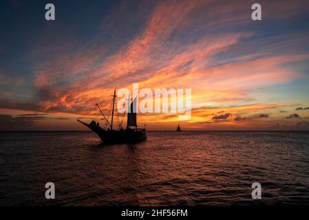 Ein feuriger Sonnenuntergang über der Karibik mit einer Piratenschifffahrt und einem Segelboot im Hintergrund. Stockfoto