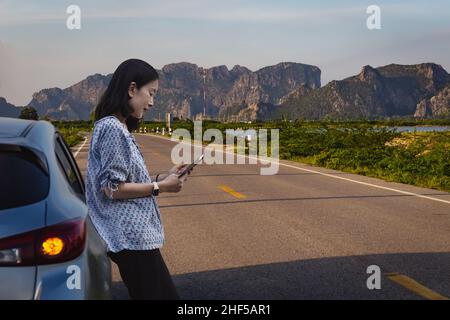 Frau, die neben dem Auto steht und versucht, das Signal auf dem Mobiltelefon zu empfangen. Stockfoto