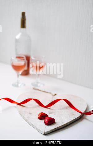 Valentinstag Festliche Tischgestaltung mit zwei Gläsern Roséwein, roten herzförmigen Schokolade Bonbons und roten Bändern auf weißem Hintergrund. Valentinstag Stockfoto