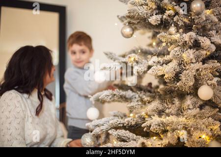 Kleiner Vorschuljunge, der Großmutter hilft, Weihnachtsbaum zu Hause zu schmücken, Großmutter und kleiner Enkel, die zusammen Weihnachtsbaum dekorieren, junge Familie Stockfoto