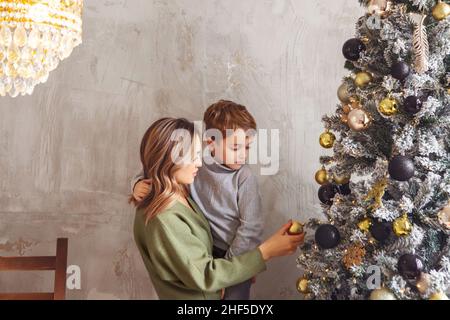 Kleiner Vorschuljunge, der Mutter hilft, Weihnachtsbaum zu Hause zu schmücken, Mutter und kleiner Sohn dekorieren Weihnachtsbaum zusammen, junge Eltern und ch Stockfoto