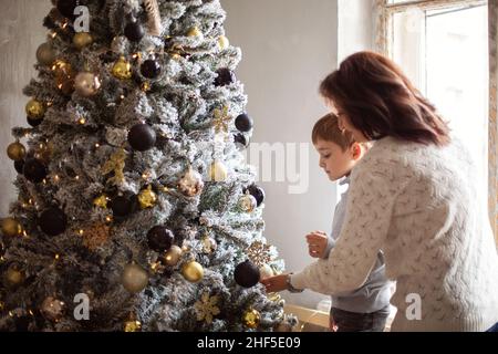 Kleiner Vorschuljunge, der Großmutter hilft, Weihnachtsbaum zu Hause zu schmücken, Großmutter und kleiner Enkel, die zusammen Weihnachtsbaum dekorieren, junge Familie Stockfoto