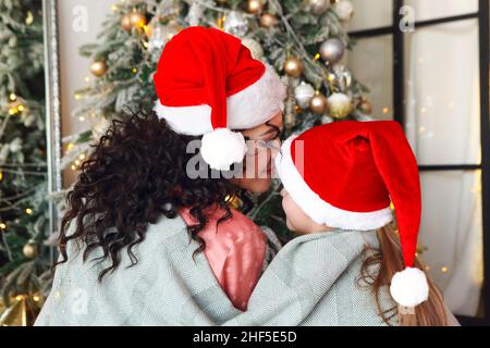 Weihnachten Für Die Ganze Familie. Rückansicht von Mutter und Kind mit weihnachtsmützen, die unter einer gestrickten Decke auf dem Boden sitzen und sich umarmen und auf den geschmückten Weihnachtsbaum blicken. Stockfoto