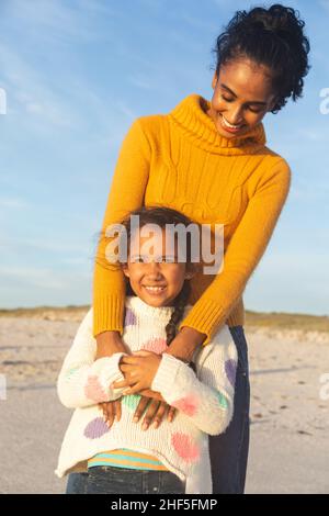 Glückliche junge Birazialfrau umarmt Tochter von hinten, während sie am Strand gegen den Himmel steht Stockfoto