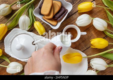 Von Hand Milch in den Kaffee in der Nähe von weißen und gelben Tulpen auf Holztisch Draufsicht gießen Stockfoto