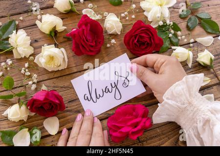 Hände mit handgeschriebener Karte, DIE IMMER von roten und cremefarbenen Rosen umgeben sind, schließen sich auf einem Holztisch aus. Femminine romantische Liebeserklärung nahe Blumen, Stockfoto