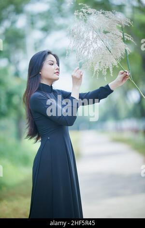 Ho Chi Minh City, Vietnam: Schönes vietnamesisches Mädchen in schwarz AO dai, posiert für ein Foto mit Unkraut Stockfoto