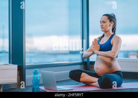 Eine Schwangerin mit namastigen Händen sitzt auf einer Matte mit einem Laptop in Lotus-Pose und praktiziert Yoga online Stockfoto