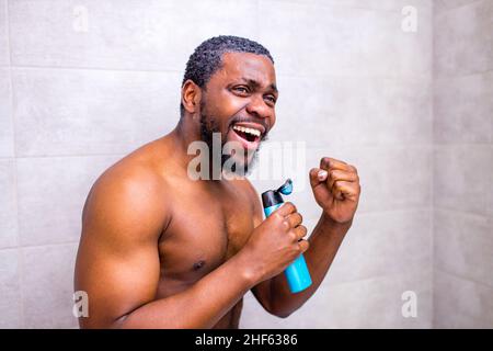 Attraktiver junger, fröhlicher Mann, der beim Waschen in der Dusche singt und die Shampoo-Flasche wie ein Mikrofon hält Stockfoto