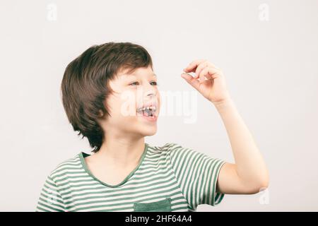 Fröhlicher Junge, der auf seinen ersten verlorenen Milchzahn unten auf grauem Hintergrund blickt. Kind zahnärztliche Gesundheitsversorgung Konzept. Stockfoto