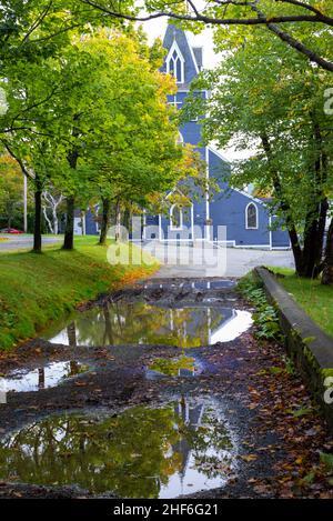 Eine alte königsblaue Holzkirche mit weißen Zierleisten, eingebettet zwischen grünen und gelben Bäumen. Es gibt eine lange Auffahrt, die zum Gebäude führt Stockfoto