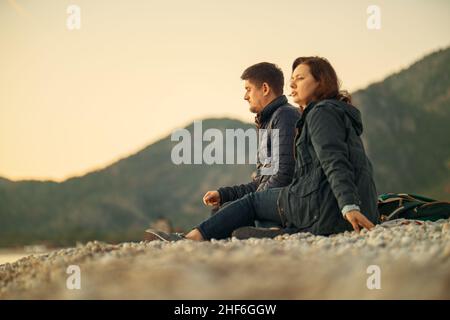 Ein Mann und eine Frau sitzen an einem Steinstrand und genießen das Wintermeer und einen wunderschönen Sonnenuntergang. Datum. Romantik. Mann und Frau. Tourismus. Reisende. Urlaub. Stockfoto