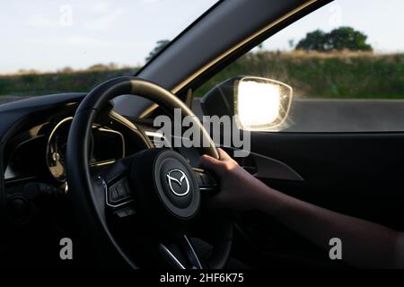 Durham, Großbritannien - 23rd. August 2019: Selbstbewusster Fahrer, der das Lenkrad eines Mazda-Fahrzeugs hält. Sonnenschein und Bäume spiegeln sich im Seitenspiegel Stockfoto