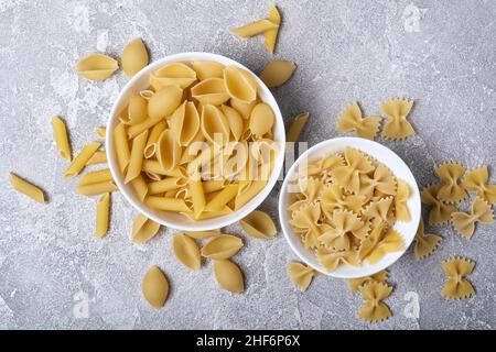 Draufsicht auf verschiedene italienische rohe Pasta in weißen Schalen auf grauem Betongrund Stockfoto