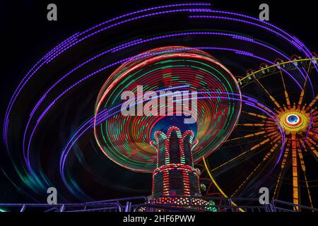 Spaß beim Karussellfahren am späten Abend, farbenfrohe Langzeitbelichtung mit leichten Spuren der Karusselldrehung mit einem Riesenrad im Hintergrund Stockfoto