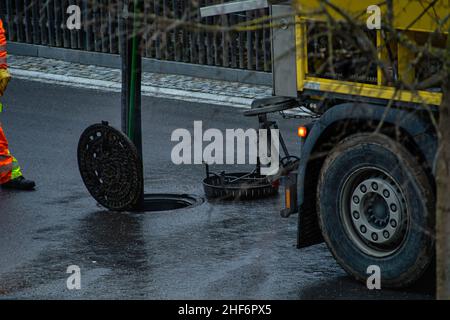 Der Abflussreinigungsarbeiter steht vor einer offenen Schachtabdeckung und erledigt seine Arbeit an einem regnerischen Tag auf einer Straße Stockfoto