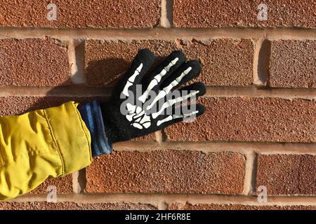 Nahaufnahme von kleinen Jungen, die Hand und Arm trugen, einen Skelett-Handschuh trugen, wobei seine Finger winkend ausgestreut an einer Ziegelwand draußen an einem sonnigen Tag aufgestellt waren. Stockfoto