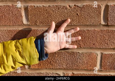 Nahaufnahme von kleinen Jungen, Hand und Arm, mit seinen Fingern, die sich an einem sonnigen Tag an einer Ziegelwand im Freien aufgestellt haben. Das kleine Kind trägt Stockfoto