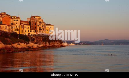 Griechenland, Griechische Inseln, Ionische Inseln, Korfu, Korfu-Stadt, sonnenaufgang am Meer, Blick auf historische Häuserzeile im Morgenlicht über dem Meer, rechts ein Schwimmer im Bild, blauer Himmel bis blassrosa Stockfoto