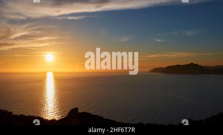 Griechenland, die griechischen Inseln, Ionische Inseln, Korfu, Westküste, Pentati, Sonnenuntergang, Blick von oben auf das Meer und Agios Gordios (im Schatten), blauer Himmel, graue Wolken, Weitwinkelansicht, Küste im Hintergrund, Sonne scheint hell knapp über dem Meer, Sonnenstrahl auf dem Wasser Stockfoto