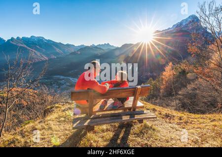 Zwei Personen (Vater und Tochter), die auf einer Bank sitzen, lachen und blicken in die Augen, während die Sonne hinter ihnen untergeht, Agordo, Provinz Belluno, Venetien, Italien Stockfoto