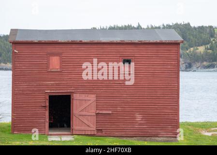 Eine alte rote Scheune aus abgenutztem und verwittertem Holz auf einer Wiese, umgeben von üppigem grünen Gras. Die Scheune hat mehrere Fenster. Hinter der Scheune ist ein Teich. Stockfoto