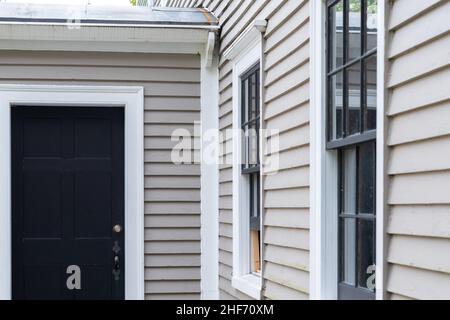 Zwei Vintage-Doppelfenster mit einer schwarzen Tür an einer beigefarbenen Holzaußenwand eines Hauses. Die Fenster sind dunkelgrün mit weißen Zierleisten. Stockfoto