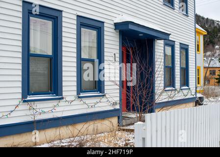 Das Äußere eines kahlen weißen Cape Cod Klappboard horizontale Holzbrett Siding Wand mit mehreren blauen Zierleisten auf doppelt hängenden Fenstern und einer roten Tür. Stockfoto