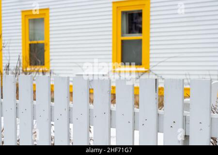 Das Äußere eines weißen Umhangs Kabeljau Klappbrett horizontale Holzbrett Stil Abstellwand mit zwei hellen gelben Zierfenstern und einem Pfostenzaun. Stockfoto