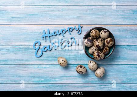 Draufsicht auf kleine Wachteleier in Schale und Text von Happy Easter auf blauem Holzhintergrund Stockfoto