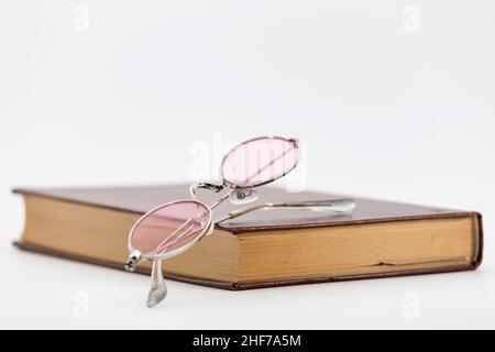 Geschlossenes altes Buch, auf das wir einige Gläser mit rosa Linsen und Metallrahmen, auf einem weißen Hintergrund platziert haben Stockfoto