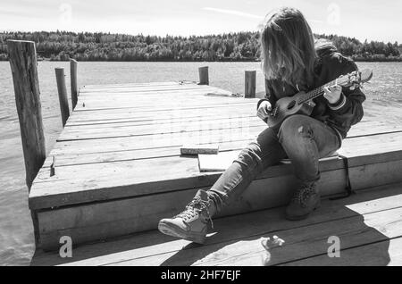 Teenager-Mädchen spielt Ukulele sitzen auf einem hölzernen Pier, schwarz-weiß-Foto im Freien Stockfoto