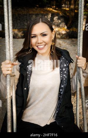 Lächelnde junge Frau auf der Schaukel im Herbst im Freien in einem warmen Pullover und ärmelloser Jacke. Stockfoto