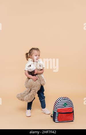 Glückliches Mädchen mit Down-Syndrom hält weiches Spielzeug, während sie in der Nähe des Rucksacks auf Beige steht Stockfoto