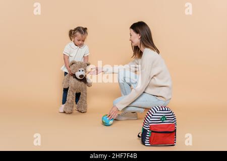 Glückliche Mutter hält kleine Kugel in der Nähe Mädchen mit Down-Syndrom umarmt Teddybär auf Beige Stockfoto