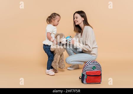 Glückliche Mutter hält kleine Kugel in der Nähe Kind mit Down-Syndrom umarmt Teddybär auf Beige Stockfoto