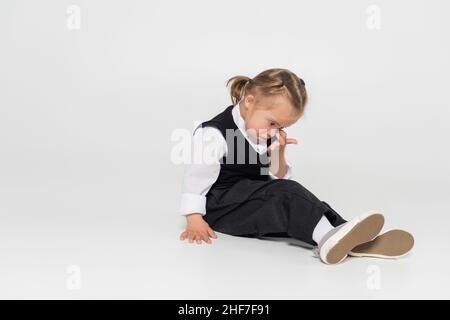 Ganze Länge des Kindes mit Down-Syndrom in Weste und weißem Hemd Reiben Auge auf grau Stockfoto