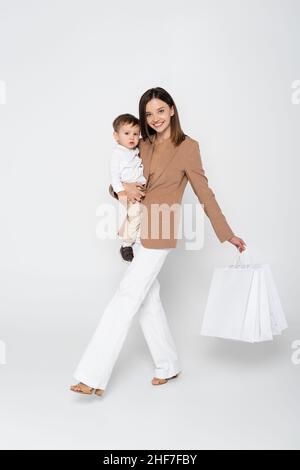 Fröhliche Mutter hält in den Armen Kleinkind Sohn und Einkaufstaschen, während auf grau zu Fuß Stockfoto
