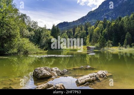 Schmölzer See bei Garmisch-Partenkirchen mit Blick auf eine Bootshütte bei gutem Wetter, Felsen im Vordergrund, große Steine ragen aus dem Wasser, Bayern, Deutschland Stockfoto