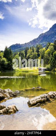 Schmölzer See bei Garmisch-Partenkirchen mit Blick auf eine Bootshütte bei gutem Wetter, Felsen im Vordergrund, große Steine ragen aus dem Wasser, Bayern, Deutschland Stockfoto