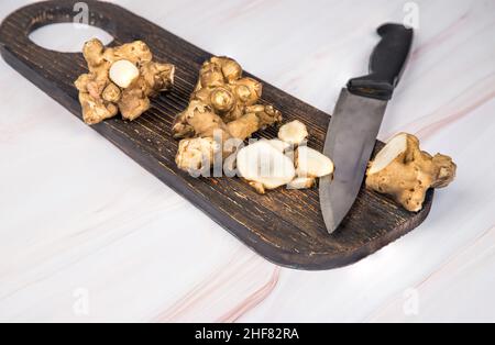 Knollen der ganzen und gehackten Jerusalem Artischocke liegen auf einem hölzernen Brett mit einem Messer auf einem hellen Tisch. Speicherplatz Kopieren Stockfoto