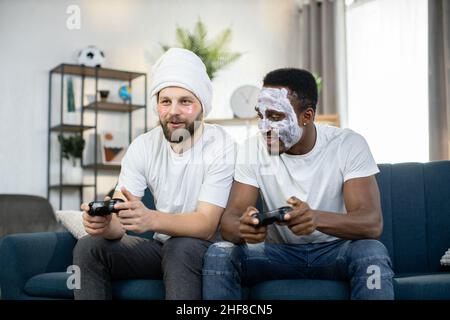 Zwei junge multiethnische Männer verbringen Freizeit zu Hause und machen Hautpflegeverfahren mit Gesichtsmaske und Augenflecken, spielen Videospiele mit Joysticks und konkurrieren miteinander Stockfoto