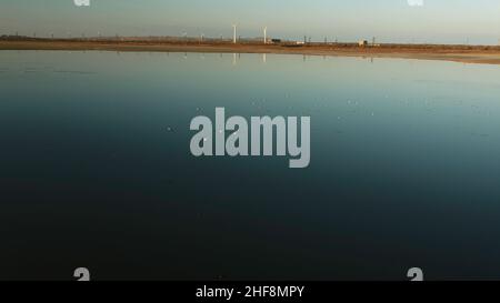 Antenne für viele Vögel, die bei Sonnenuntergang über dem See fliegen. Eine silhouettierte Herde wilder Enten, die über dem dunkelblauen Wasser vor der strahlenden Sonne und dem blauen Himmel emporragen. Stockfoto