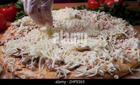 Sauce hinzufügen. Italienische Pizzazubereitung. Rahmen. Käse wird auf Tomatensauce auf Pizzabasis verteilt. Nahaufnahme Hand des Chefbäckers in weißer Uniform Herstellung Stockfoto