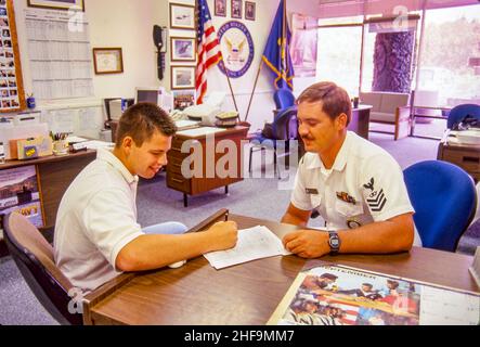 In einem Rekrutierungsbüro in Fullerton, CA, meldet sich ein junger Mann bei der US-Marine an, während der uniformierte Rekrutierer zuschaut. US-Flagge beachten. Stockfoto